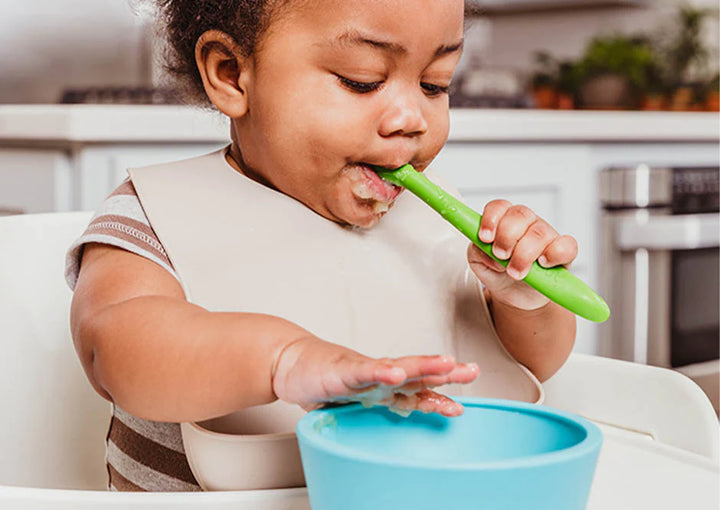 baby eats out of a blue silicone suction bowl with a green silicone baby spoon while wearing a off white silicone bib
