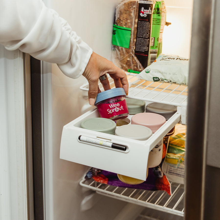 Woman holding a a jar of puree with blue top out of glass jar storage tray in freezer with other multi colored jars in the tray