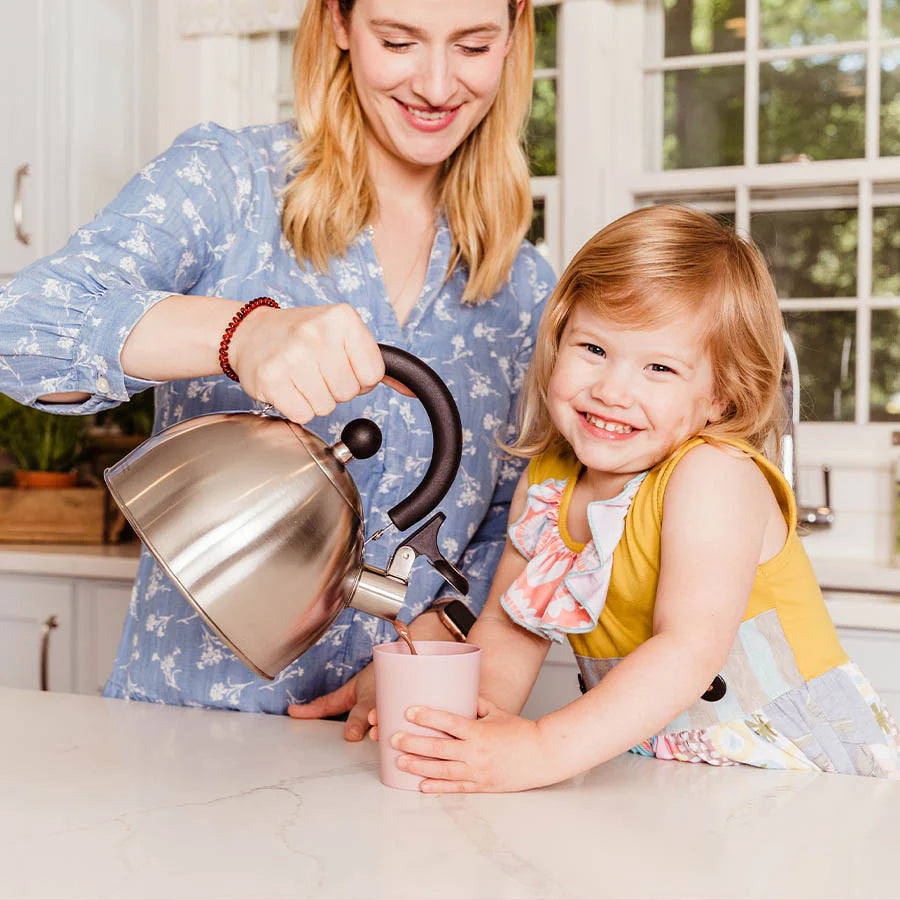 Mom helps child pour a liquid into pink bamboo cup in kitchen