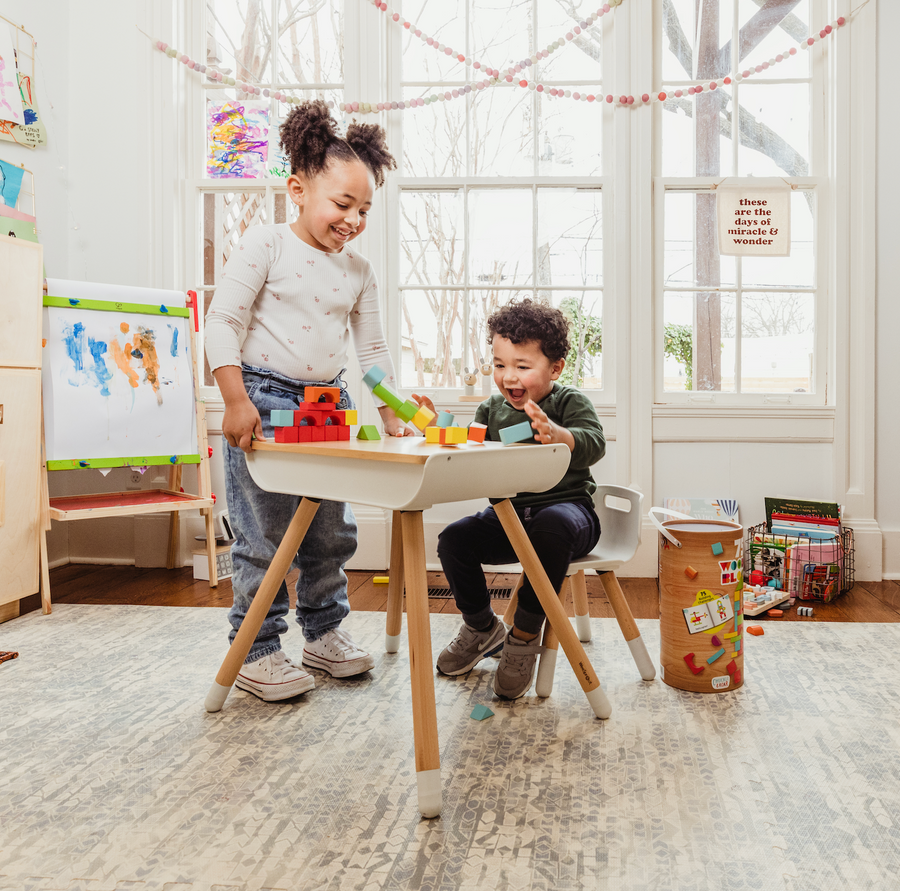 Two children play with blocks at a kids desk and chair set in front of a window