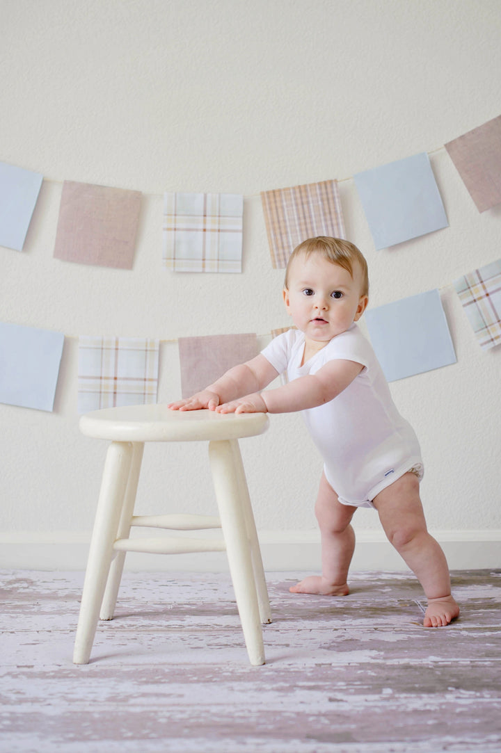 Baby standing next to stool on carpet with a blue and pink banner garland on the wall behind. Baby is wearing a white onsie. 