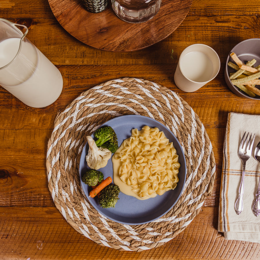Pasta and veggie meal on blue bamboo toddler plate with placemat under it on a wooden table with milk and silverware.