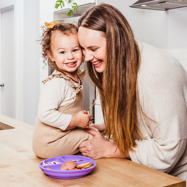 Mom hugs kid sitting on counter with food in purple silicone divided plate on counter