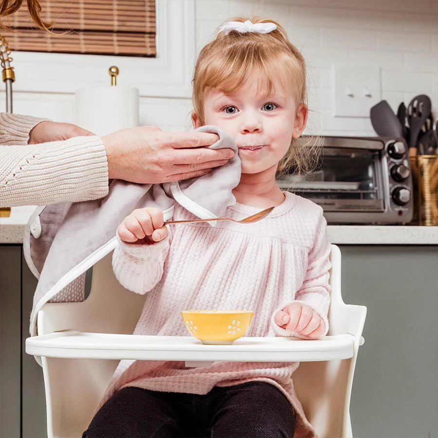 Mom cleans her toddler face while sitting in high chair eating with spoon