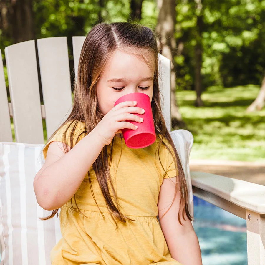Toddler sips drink out of red bamboo cup while sitting in chair outside