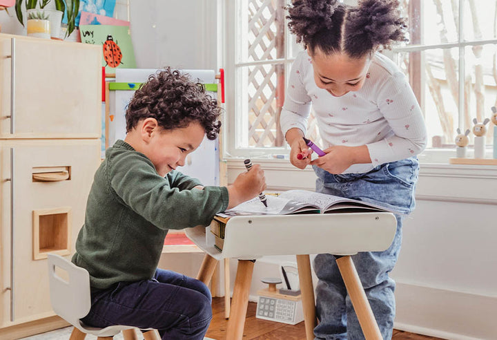 Two kids play with markers and coloring book on a white kids desk and chair set