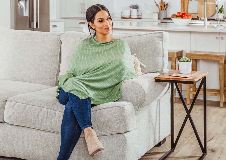 Mom sits on couch in green nursing pancho while breastfeeding a baby in her home.