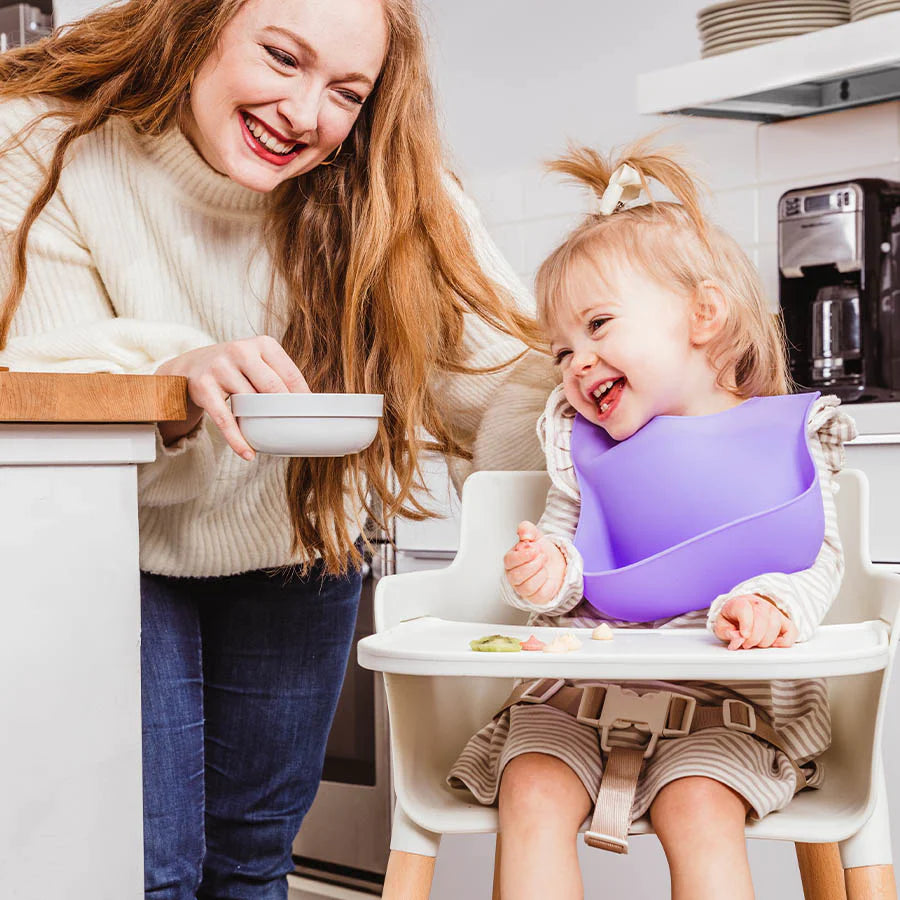 Mom brings food in bowl to toddler sitting in high chair in a purple silicone bib