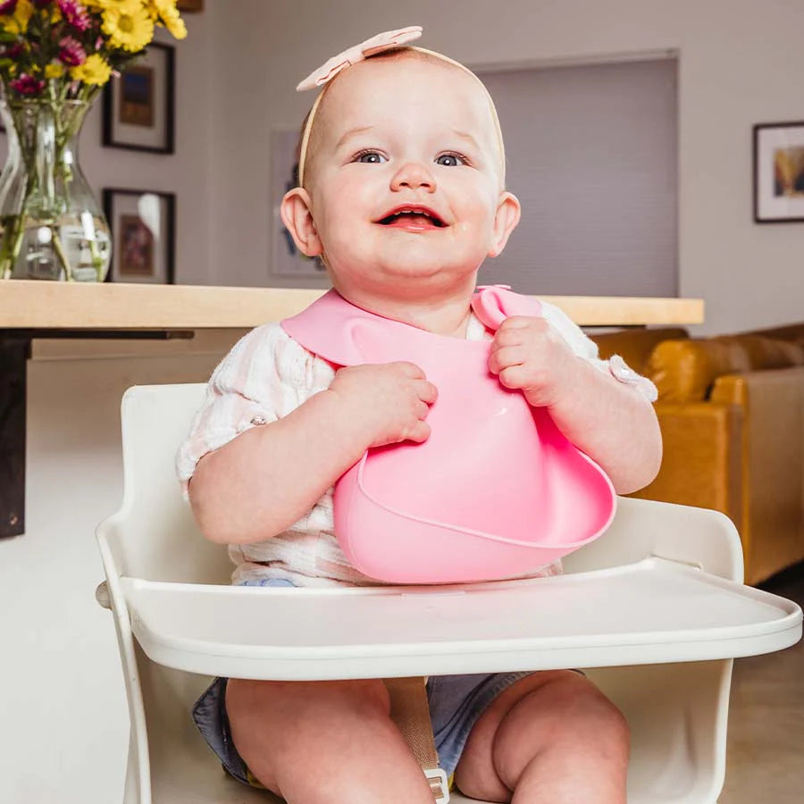 baby sits in high chair while wearing a pink silicone bib