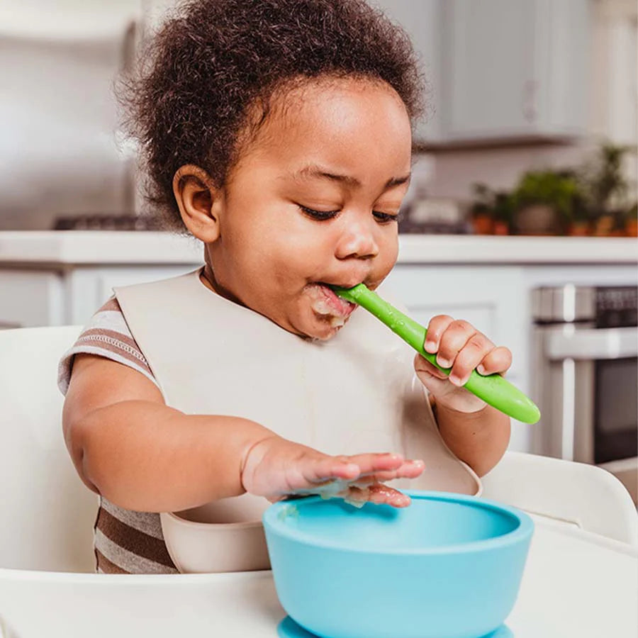Baby eats food in high chair out of a blue silicone bowl and green silicone baby spoon in white silicone bib
