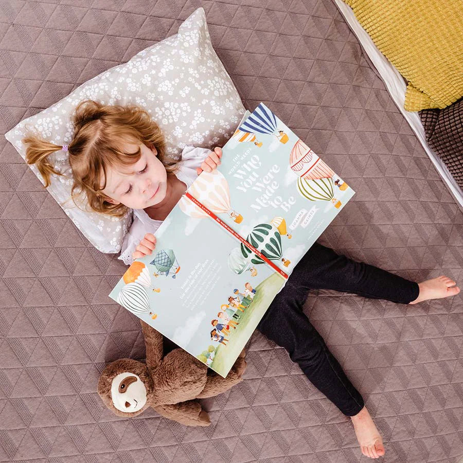 Child lays in bed with a book on a toddler pillow