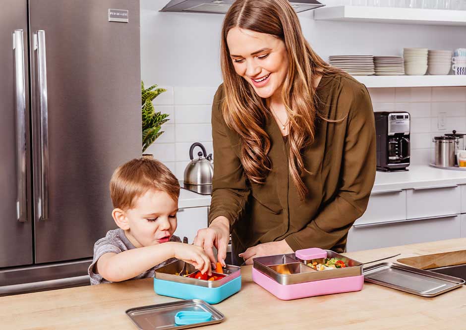 Mom and son pack lunches in a blue small stainless steel bento box with sleeve and pink large stainless steel bento box with sleeve in kitchen